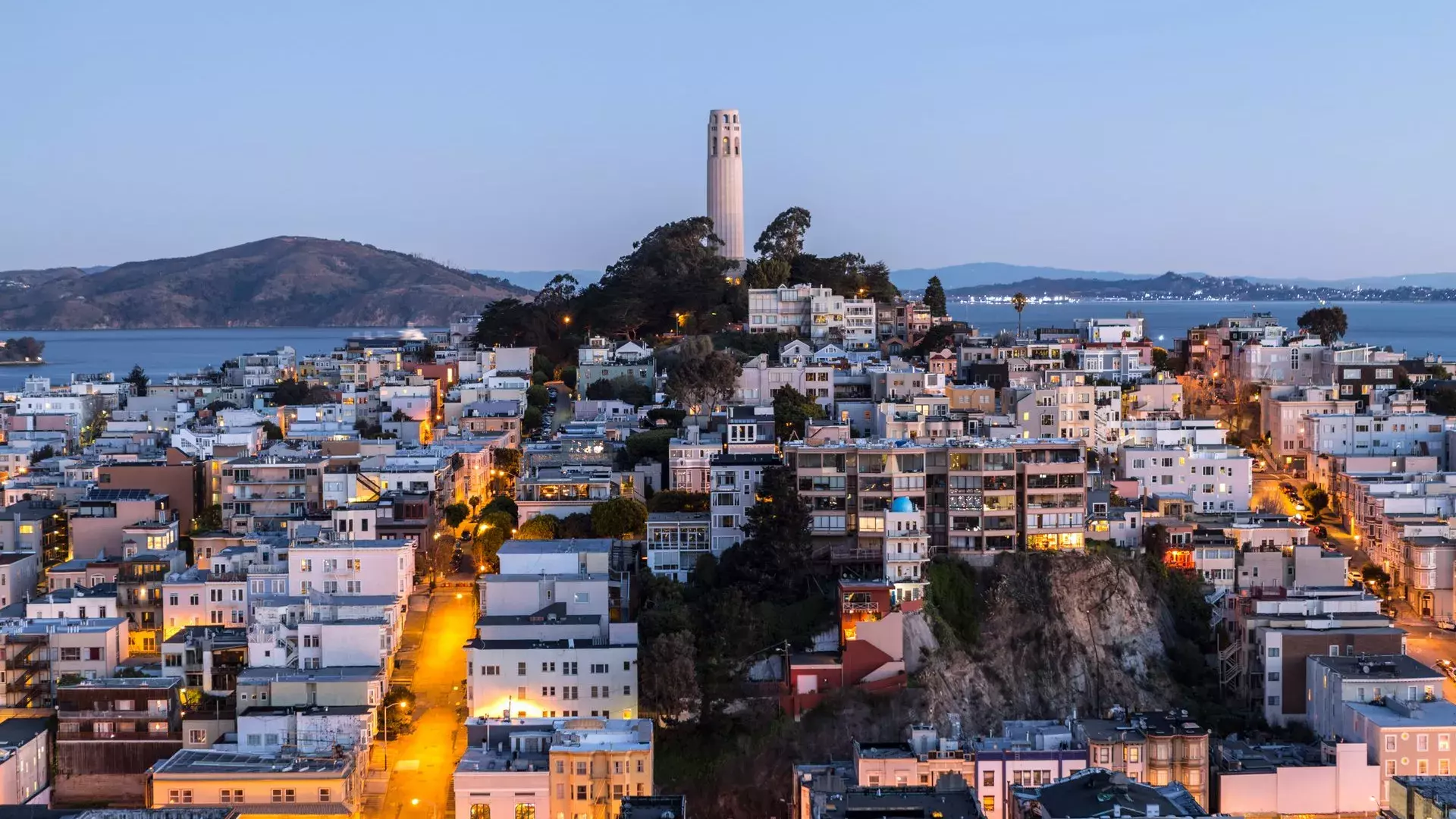 황혼 무렵의 샌프란시스코 Coit Tower 타워 앞에는 조명이 켜진 거리가 있고, 타워 뒤로는 샌프란시스코 만이 보입니다.